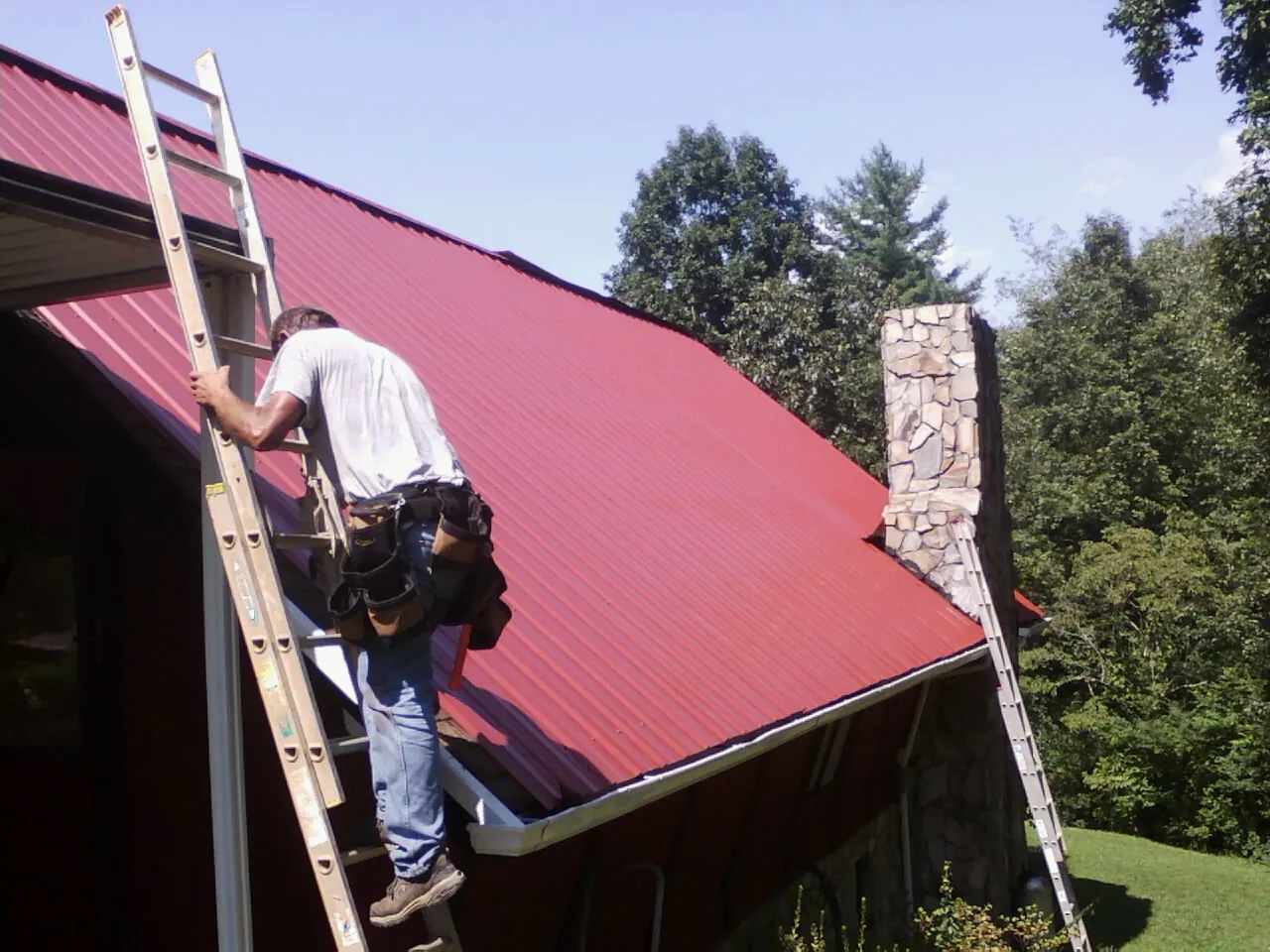 Roofer on a ladder beside a red metal roof and stone chimney — Soffit & Fascia Repair by Roofing Brothers in Davis City